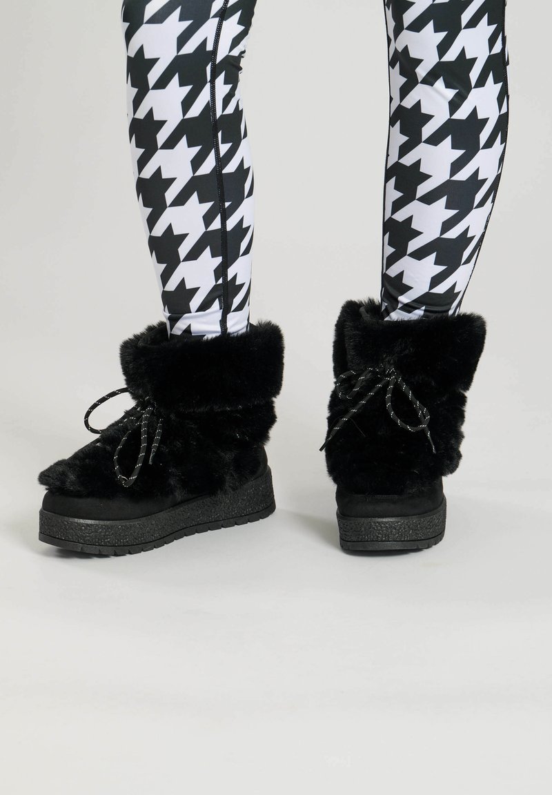 Black fur-lined boots with rounded toes, flat textured soles, and bow accents on the shaft, worn with black and white houndstooth leggings.