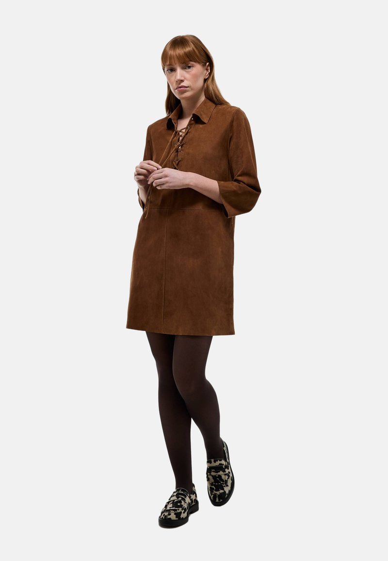 Robe en daim marron avec col, lacets à l'avant et manches aux trois quarts. Le mannequin porte des collants marron et des chaussures à motifs. Design simple et droit.