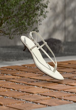 Sandale blanche à talon kitten avec des lanières et un bout pointu, flottant au-dessus d'un sol en carrelage en bois en plein air avec un arrière-plan flou de plante et de mur.