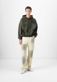 Olivengrønn hettegenser med "ATELIER" trykk, paret med kremfargede joggebukser med en falmet svart gradienteffekt og hvite sneakers.