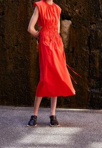 Person som bär en ärmlös ljusröd midiklänning med sidouttag och svarta öppna vävda skor står på en strukturerad mark.