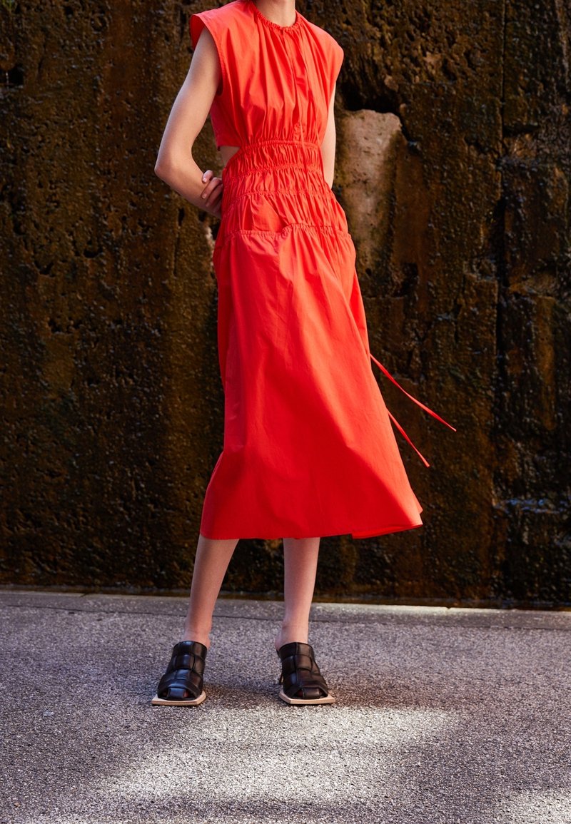 Person som bär en ärmlös ljusröd midiklänning med sidouttag och svarta öppna vävda skor står på en strukturerad mark.