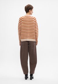 Maglione a righe in beige e arancione con un fit rilassato, abbinato a pantaloni marroni larghi. L'outfit presenta polsini a costine e stivali alti fino alla caviglia.