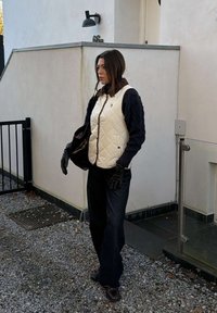 Kvinde står på grus uden for en bygning iført en hvid quiltet vest, sort sweater, vide jeans, sorte handsker og bærer en stor sort taske.