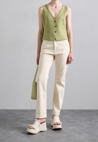 Ljusgrön ärmlös väst med tre framknappar, tillsammans med beiga rakbenbyxor och krämfärgade plattformssandaler med fransdetaljer.