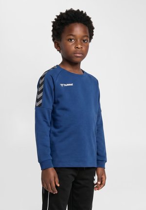 Ung dreng med krøllet hår iført en blå langærmet sportsshirt og sorte bukser, stående mod en ensfarvet lys baggrund.