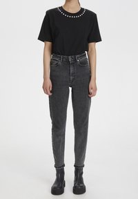 Camiseta negra con acentos decorativos de perlas alrededor del escote, combinada con jeans negros de talle alto y desgastados, y botas negras gruesas.