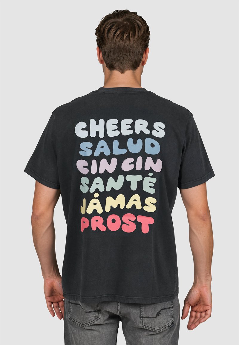 Homme portant un T-shirt noir avec les mots colorés « Cheers », « Salud », « Cin Cin », « Santé », « Jámas » et « Prost » imprimés au dos.