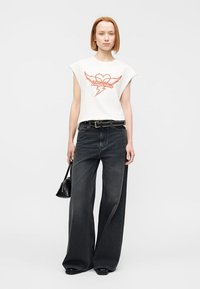 Γυναίκα με κοντά κόκκινα μαλλιά, φορώντας λευκό αμάνικο τοπ με γραφικό κόκκινης καρδιάς και αστραπής, φαρδιά μαύρα τζιν, μαύρη ζώνη, κρατώντας μικρή μαύρη τσάντα.