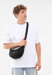 Borsa a spalla nera con una forma curvata, realizzata in tessuto, con una piccola etichetta frontale, indossata da una persona in una semplice maglietta bianca.