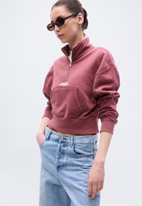 Μπλούζα τύπου sweatshirt σε μωβ σκούρο χρώμα με φερμουάρ μέχρι τη μέση και μπροστινή τσέπη, από μαλακό ύφασμα και με ριμπ μανσέτες, συνδυασμένη με ανοιχτό μπλε τζιν ψηλόμεσο.