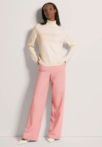 Beige coltrui met geribbelde manchetten, gecombineerd met losse roze broek. Model staat tegen een neutrale achtergrond en draagt witte hakken.