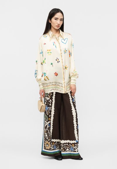 Γυναίκα που φοράει κρεμ μπλούζα με πολύχρωμα αφηρημένα σχέδια, φαρδιά μαύρη παντελόνα με λουλουδένια μοτίβα, κρατώντας μπεζ τσάντα χειρός.