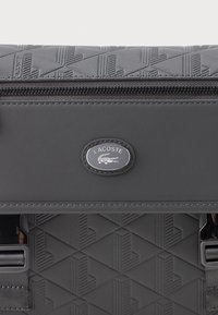 Bolsa Lacoste cinza-escuro com padrão geométrico em relevo, emblema prateado, fecho de zíper e fechos de alça pretos, apresentada em close-up.