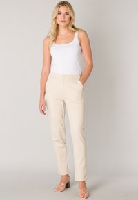 Beige slimfit broek met zijzakken en knoopsluiting, gecombineerd met een witte tanktop. Glad materiaal en taps toelopende pijpen.