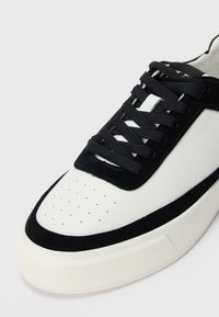 Witte sneaker met zwarte suède details, zwarte veters, geperforeerde neus en dikke witte zool tegen een effen lichte achtergrond.