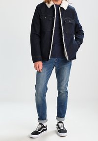 Tmavě modrá džínová bunda s bílým umělým beránkem na límci, dvě kapsy na hrudi. Nošena přes námořnickou košili, doplněná světle modrými džínami a černými teniskami.