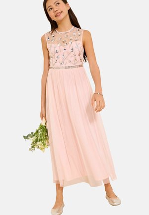 Mädchen in einem blassrosa ärmellosen Kleid mit floralem Schmuck hält einen kleinen Strauß weißer Blumen und trägt passende Ballettschuhe.