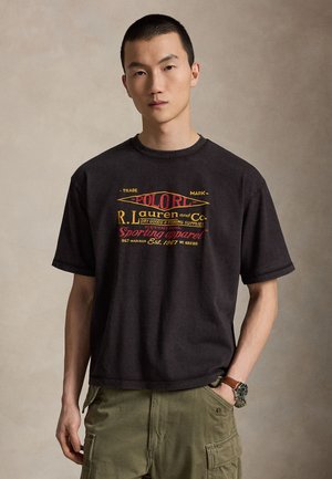 Ung mand med kort hår iført en sort grafisk t-shirt og olivengrønne cargobukser, stående med den ene hånd i lommen.
