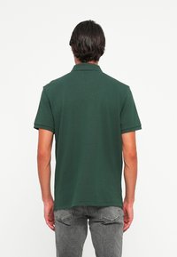 Man in een donkergroen polo met korte mouwen en grijze jeans, staande met de rug naar de camera tegen een effen witte achtergrond.