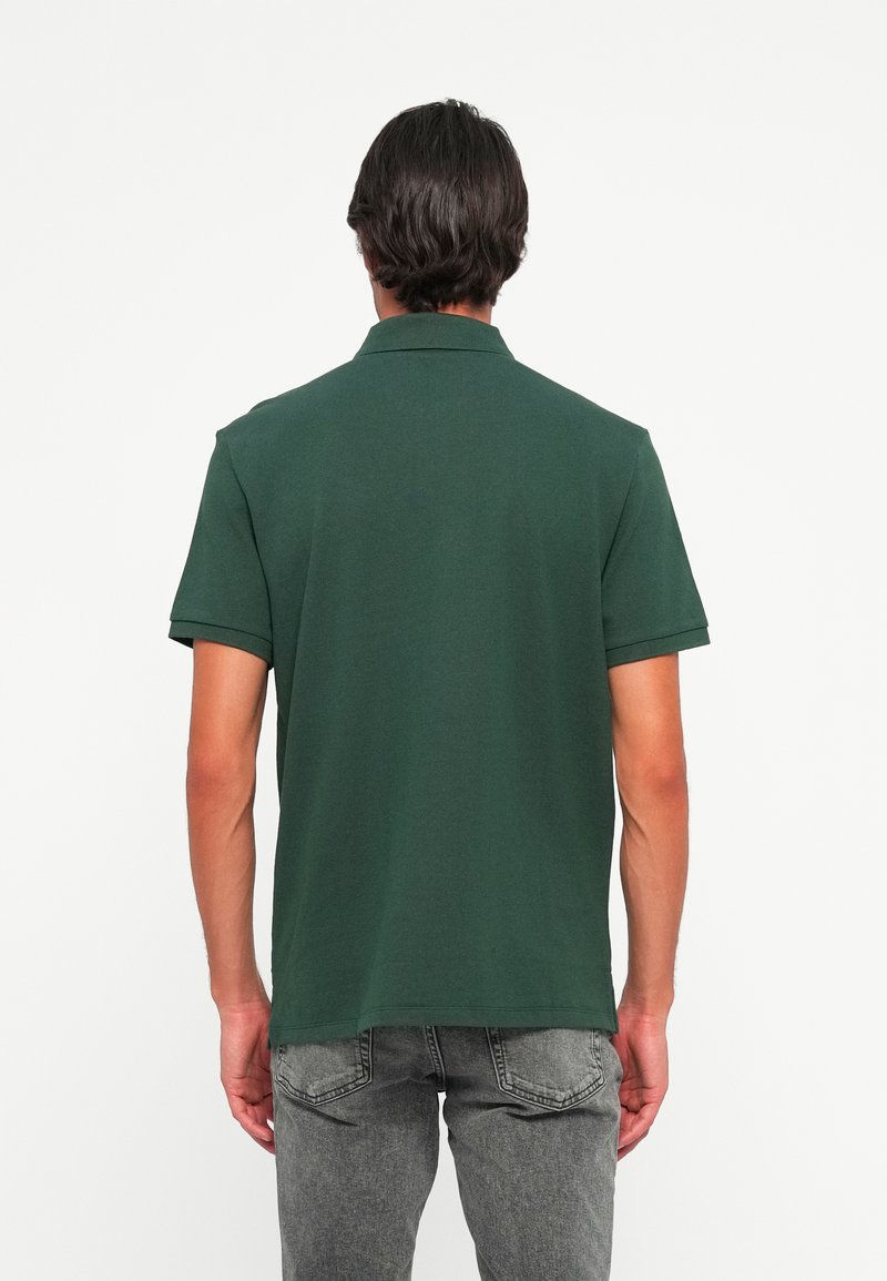 Mężczyzna w ciemnozielonej koszulce polo z krótkim rękawem i szarych dżinsach, stojący tyłem na jednolitym białym tle.