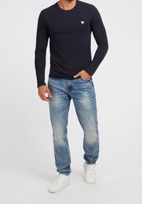 Marinblå långärmad T-shirt med en liten triangulär logotyp i kombination med ljusblå slitna jeans och vita sneakers. Avslappnad, figurnära silhuett.