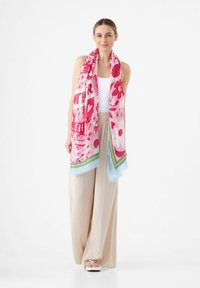 Roze bloemen sjaal met gedurfde patronen en lichtblauwe accenten, gemaakt van zachte stof, over de schouders gedrapeerd. Gecombineerd met lichtbeige broeken.