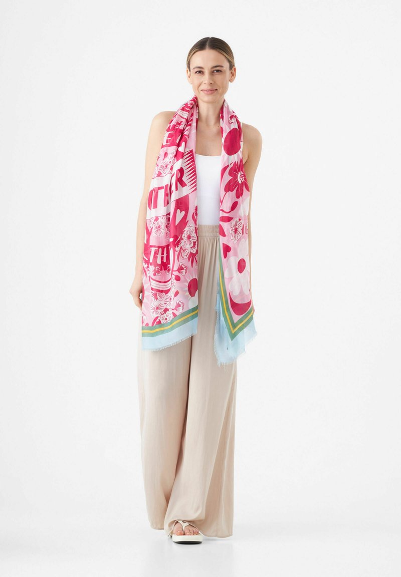 Roze bloemen sjaal met gedurfde patronen en lichtblauwe accenten, gemaakt van zachte stof, over de schouders gedrapeerd. Gecombineerd met lichtbeige broeken.