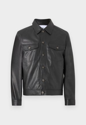 Mladi muškarac stoji, nosi crnu kožnu jaknu, bež pulover, sive široke hlače i crne mokasinke na bijeloj pozadini.