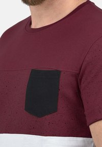 Kortærmet maroon T-shirt med en sort lomme og en hvid nederste sektion. Stoffet har et tekstureret, spraglet mønster på den øverste del.