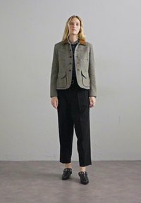 Jakna iz volne s houndstooth vzorcem in rjavim usnjenim ovratnikom, z dvema sprednjima žepoma, v kombinaciji s širokimi črnimi hlačami in črnimi čevlji s kovinskimi dodatki.