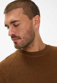 Bruine gebreide trui met een gestructureerd oppervlak en een ribbelachtige ronde hals, getoond op een model dat naar de zijkant kijkt.