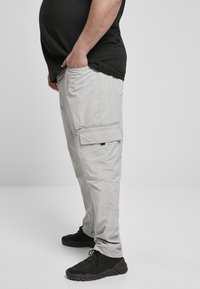 Hellegraue Cargo-Hosen mit Taschen, aus leichtem Material. Kombiniert mit schwarzen Sneakers und einem schwarzen Shirt. Seitenansicht.