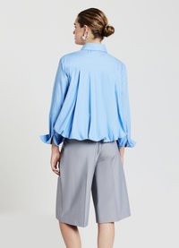 Hellblaue Langarmbluse mit geschwungenem Saum, Rückenfalten und geknöpften Manschetten, kombiniert mit grauen, knielangen, maßgeschneiderten Shorts.