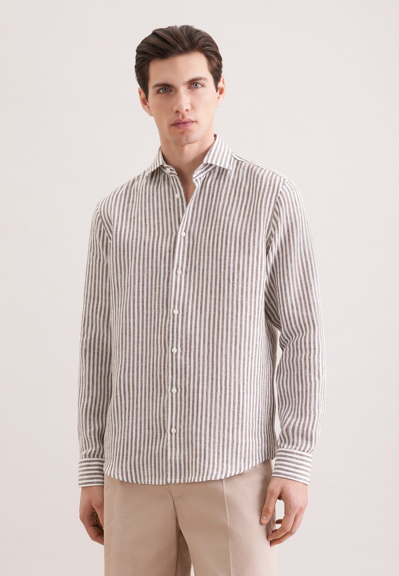 Homme portant une chemise à rayures verticales beige et blanche à boutons et un pantalon beige clair, debout devant un fond clair uni.