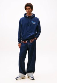 Navy blue hoodie with "Tommy" logo, layered over a plaid shirt. Paired with loose-fitting dark denim jeans and unique mixed-texture sneakers.