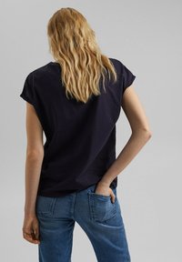 Marineblå bomulds t-shirt med korte opsmøgede ærmer, vist fra ryggen. Parret med lyseblå denimjeans med en baglomme.