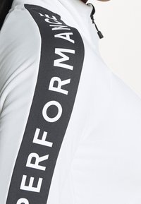 Witte hoge nek top met een zwarte verticale streep waarin het woord "PERFORMANCE" in vette witte letters staat. Glad stoftextuur.