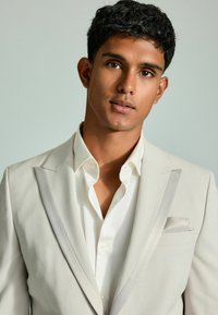 Hellbeige Anzugjacke mit Schalkragen, kombiniert mit einem weißen Hemd. Verfügt über ein Einstecktuch und eine glatte Stoffstruktur. Keine Muster sichtbar.