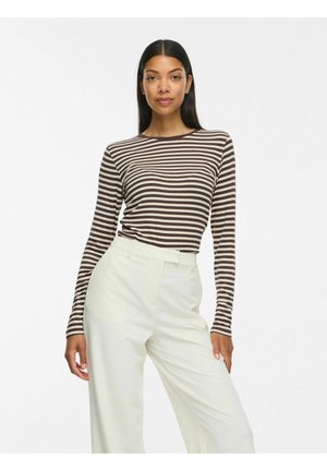 Femme aux longs cheveux foncés portant un haut à manches longues rayé marron et blanc et un pantalon blanc taille haute à jambes larges, debout devant un fond blanc.