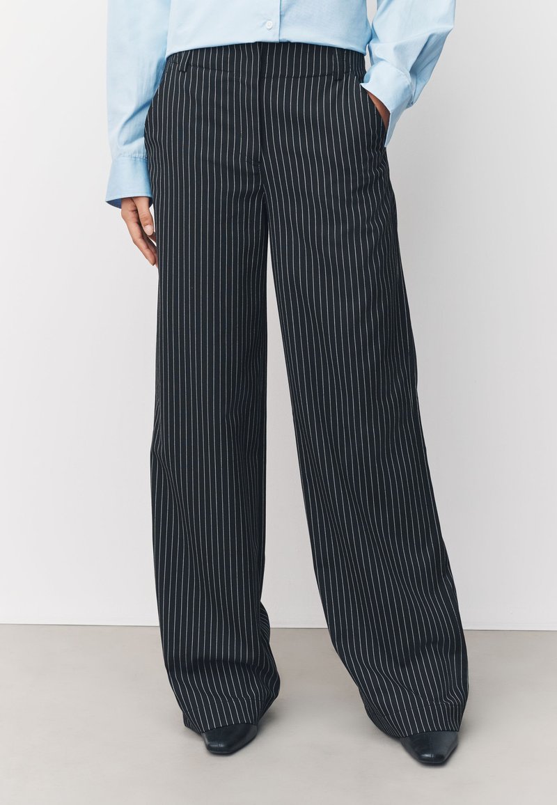 Person trägt schwarze, weit geschnittene Nadelstreifenhose und ein hellblaues Langarmhemd, steht mit einer Hand in der Tasche.