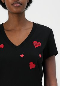 Schwarzes V-Ausschnitt-T-Shirt mit roten gestickten Herzen in verschiedenen Größen, aus weichem, atmungsaktivem Material mit kurzen Ärmeln.