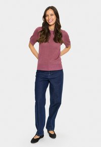 Lila, kurzärmelige, strukturierte Strickjacke mit rundem Halsausschnitt, kombiniert mit lässigen, locker sitzenden blauen Jeans und schwarzen flachen Schuhen.