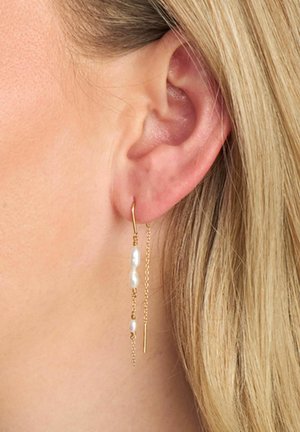 Øreringe i guldfarve med kædedesign, der har en stang og to hvide perleaccents, vist på en models øre.