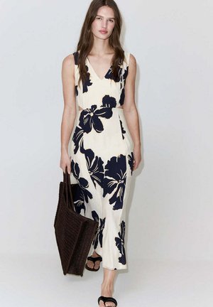 Mujer con vestido blanco sin mangas con un gran estampado floral negro, lleva un bolso tote tejido de color marrón oscuro y sandalias negras.