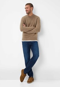 Brązowy sweter dzianinowy o ribbowanej fakturze, w połączeniu z niebieskimi dżinsami i brązowymi zamszowymi butami. Model pozuje z rękami skrzyżowanymi na tle białej ściany.