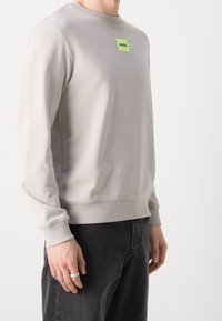 Lichtgrijze sweatshirt met geribbelde manchetten en zoom, voorzien van een klein fluorescerend groen vierkant logo met de tekst "HUGO" op de borst. Zachte stoftextuur.