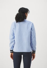 Ljusblå fleecejacka med hög krage, texturerad yta och ribbade ärmslut. Bärs över mörka leggings med logotypdetaljer.