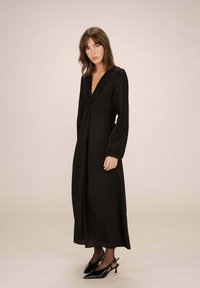 Robe noire à manches longues avec un décolleté en V plongeant, taille froncée et jupe fluide. Associée à des escarpins à brides arrière noirs. Texture lisse.