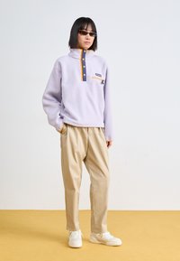 Ανοιχτό μωβ φλις πουλόβερ με τσέπη στο στήθος με φερμουάρ, πορτοκαλί λεπτομέρειες και ναυτικές κουμπιά. Συνδυασμένο με κρεμ χρώματος παντελόνι σε χ�αλαρή γραμμή.